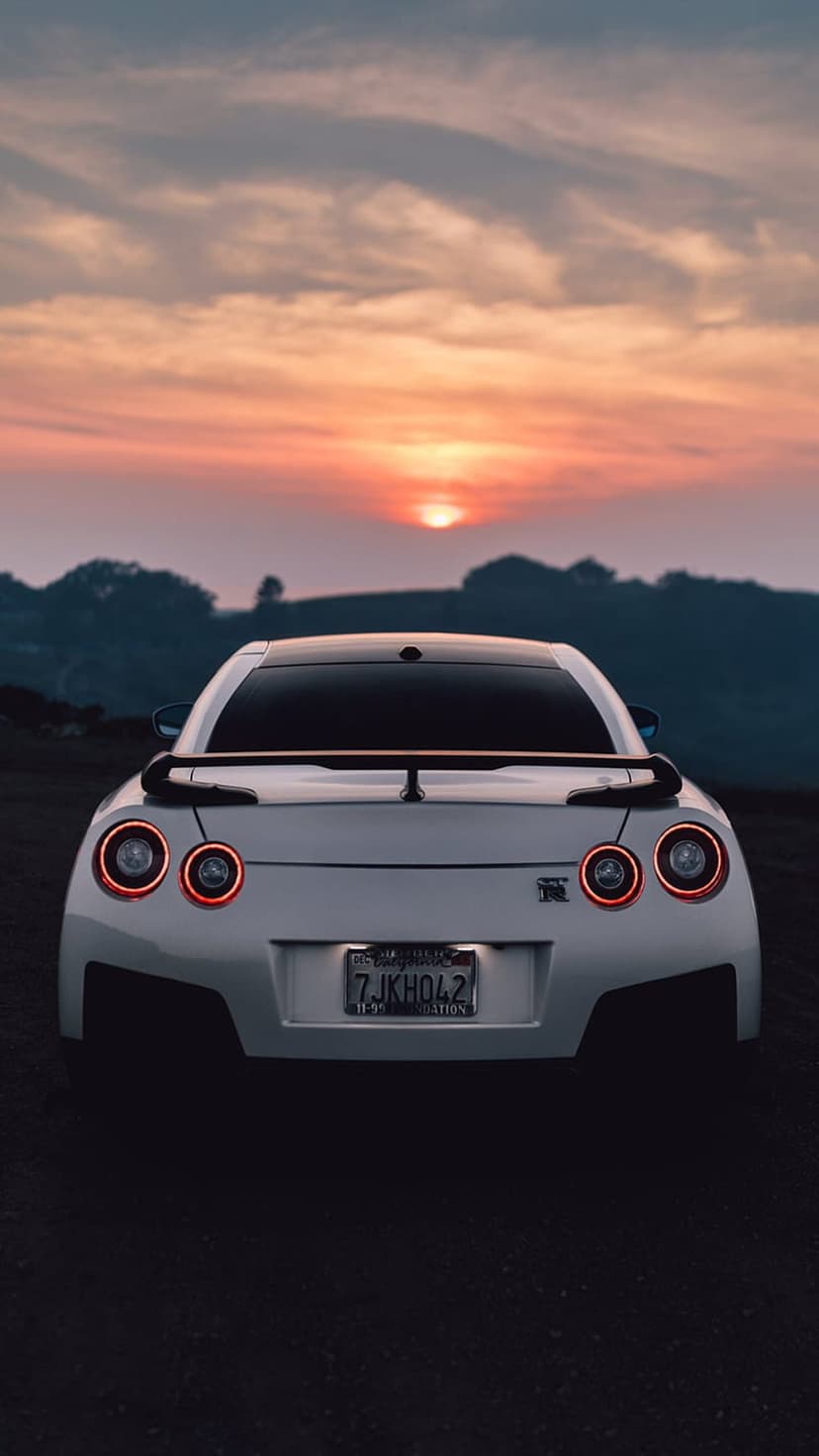 White Nissan GT-R parked on dark rocks with illuminated red taillights under a pink and orange sunset sky.