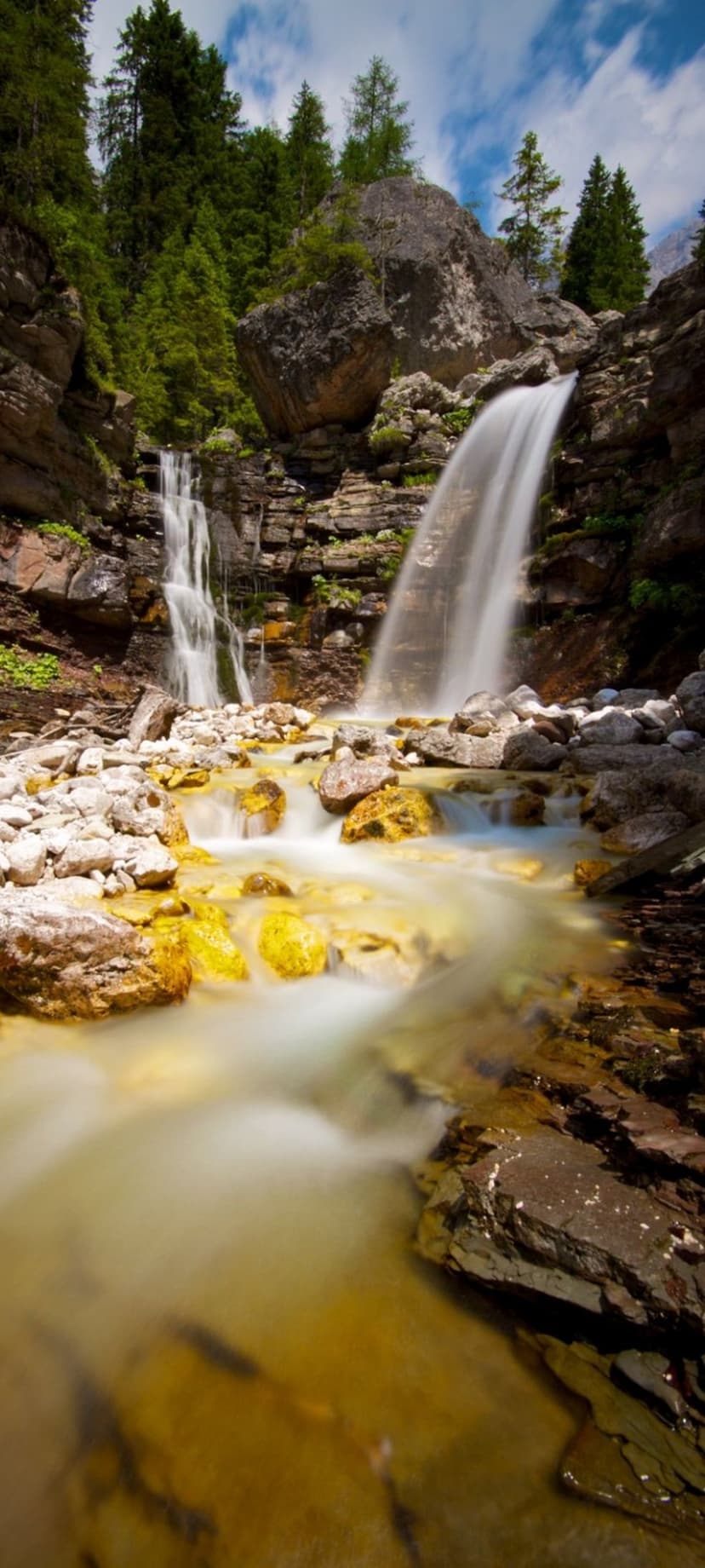 Waterfall cascading over dark cliffs into a golden rocky stream surrounded by dense green trees and soft sunlight.