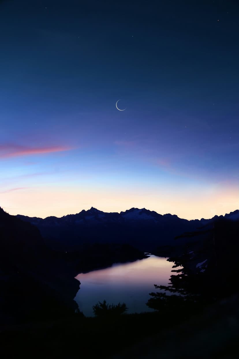Slender crescent moon and stars above jagged mountain silhouettes reflecting in a calm alpine lake at twilight.