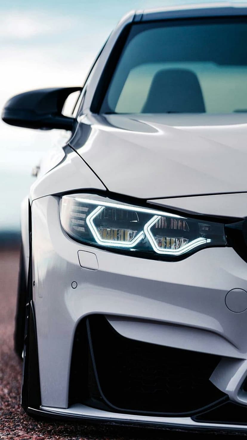BMW M4 headlight glows with illuminated LED rings against a white car body and blurred asphalt road background.