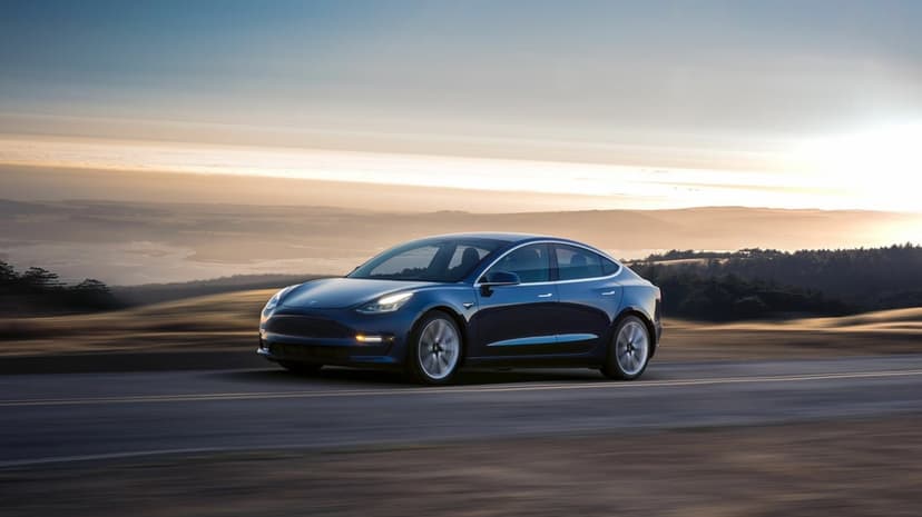 Blue Tesla Model 3 driving on a coastal road during a hazy golden hour sunset with rolling hills in the distance.