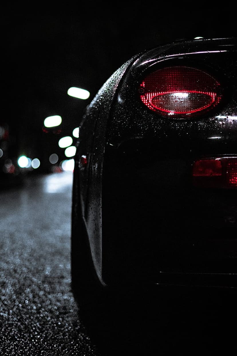 Red car tail light glowing on a dark wet surface with glistening water droplets and blurred bokeh street lights.