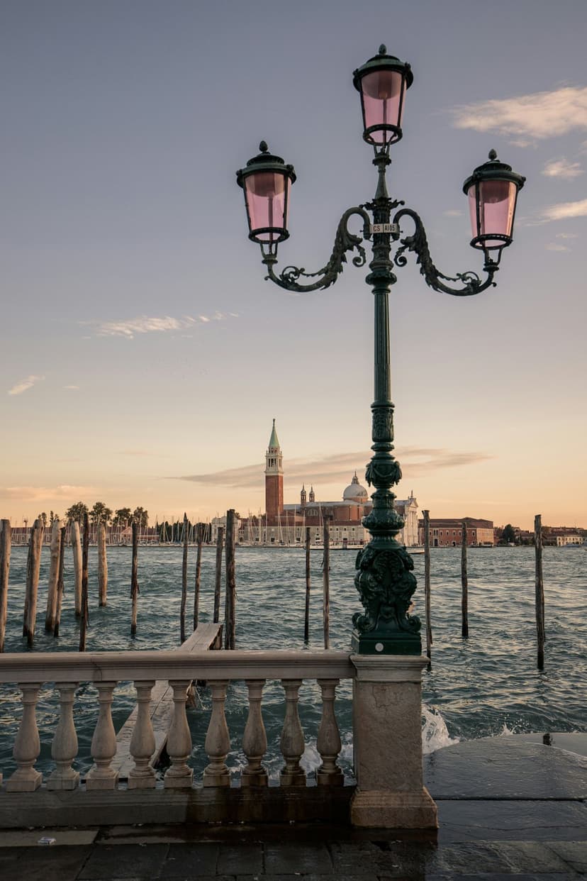 Ornate green lamppost with pink glass stands before a Venetian canal and bell tower under a sunset sky.