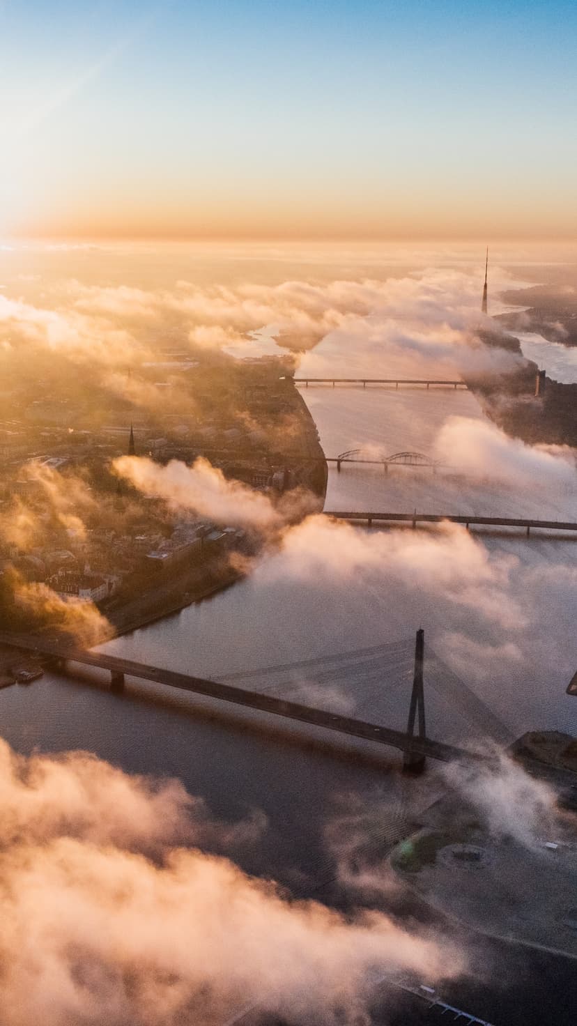 Riga bridges pierce through thick white morning mist under a glowing golden sunrise sky from an aerial view.