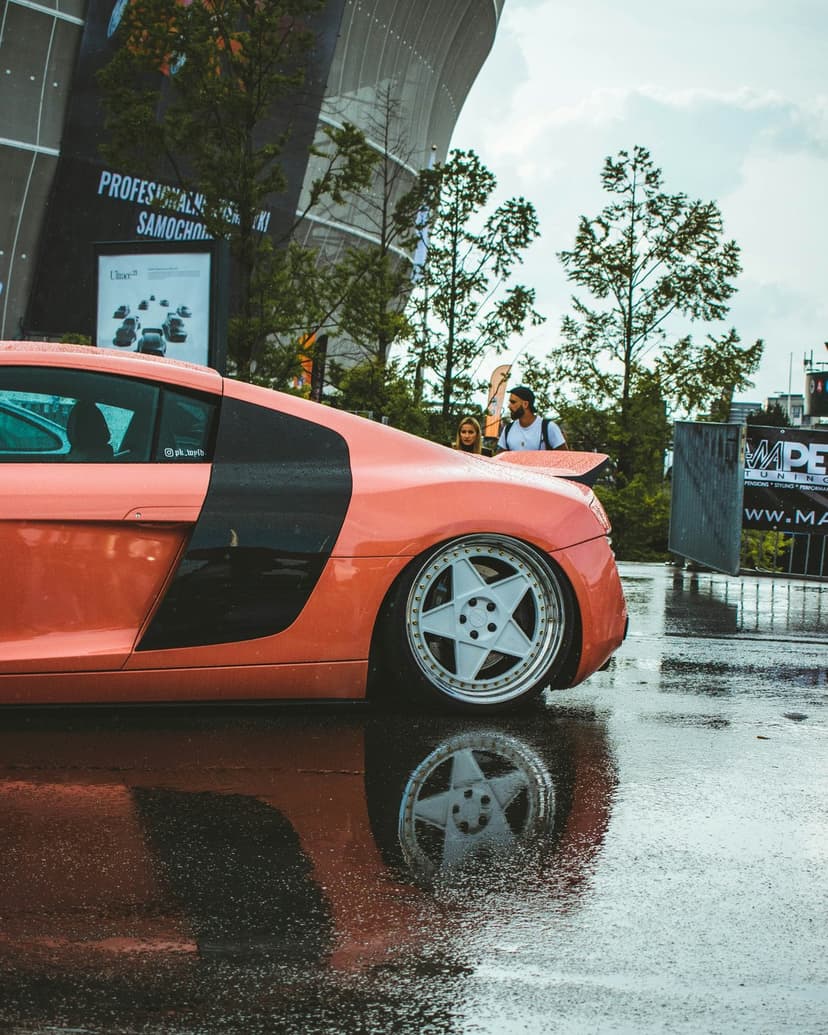 Orange sports car parked on wet asphalt with a clear reflection and detailed black custom wheels after rain.