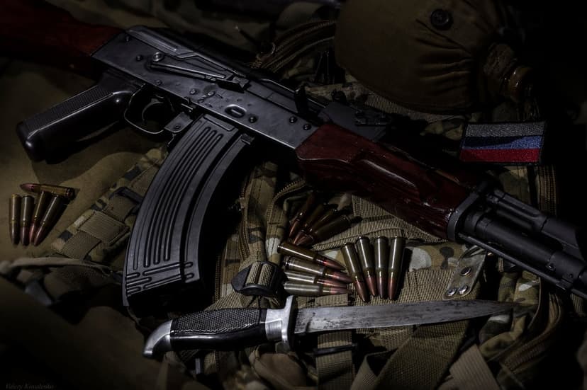 AK-47 assault rifle with a combat knife and brass ammunition resting on a camouflage surface with a flag patch.
