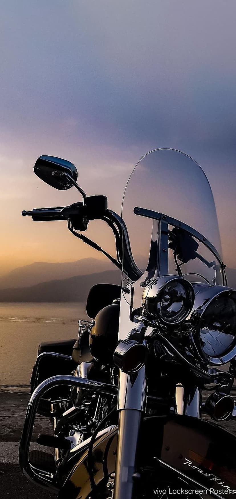 Chrome motorcycle parked by a calm lake during a vibrant purple and orange sunset with reflections on the water.