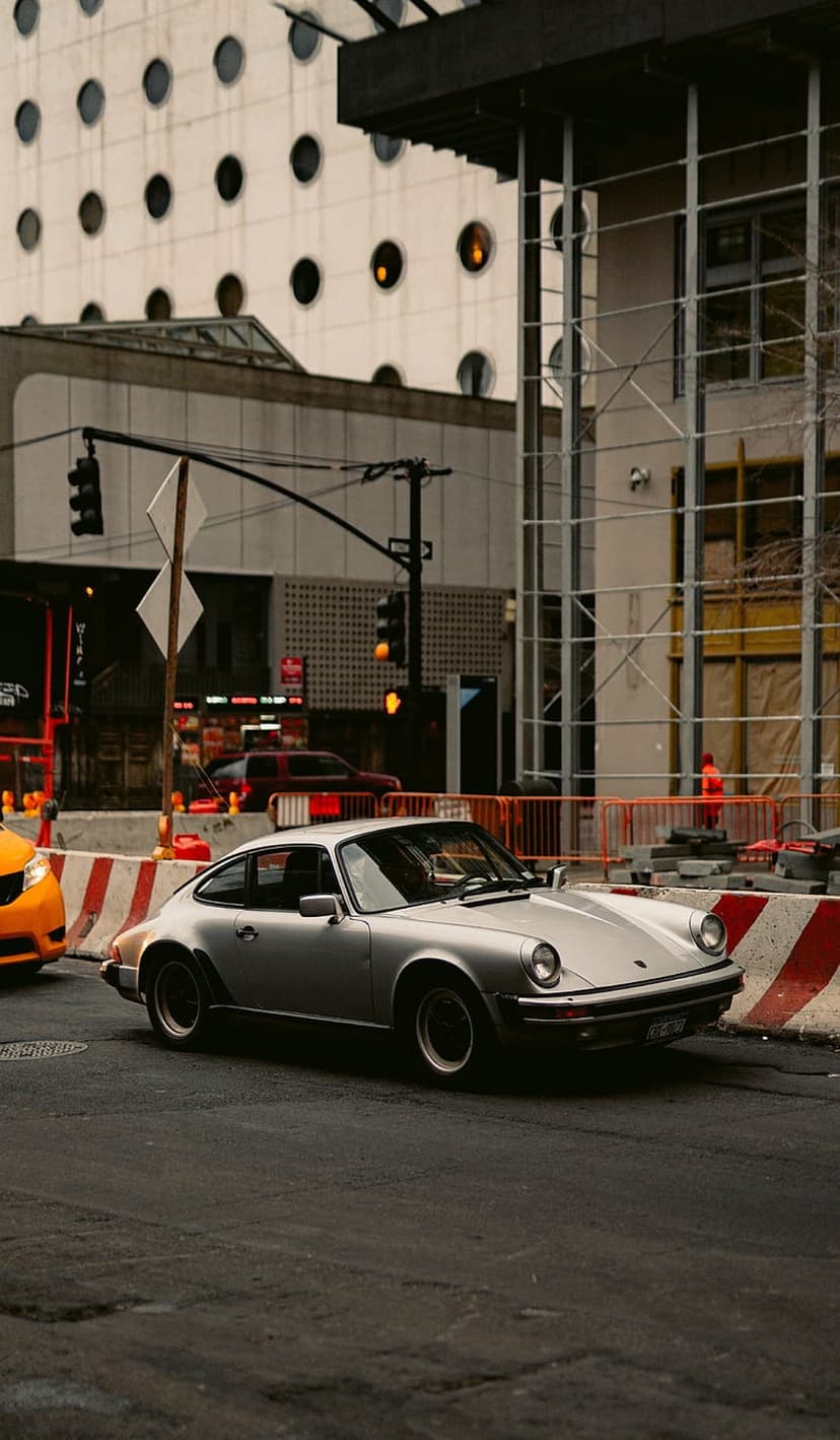 Silver Porsche 911 drives on a city street past orange construction barriers and scaffolding under a tall building.