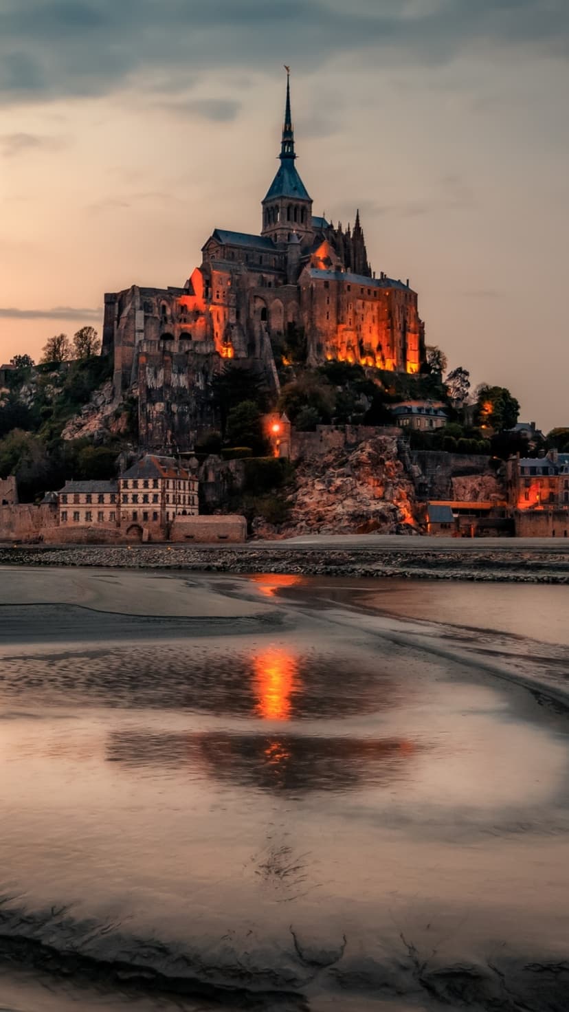 Mont Saint-Michel abbey towers over calm tidal flats and water under a soft purple and gold twilight sky.