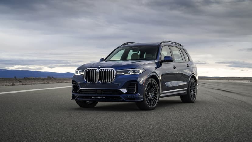 Blue Alpina XB7 luxury SUV parked on a wide asphalt runway under a dramatic grey cloudy sky with mountains.