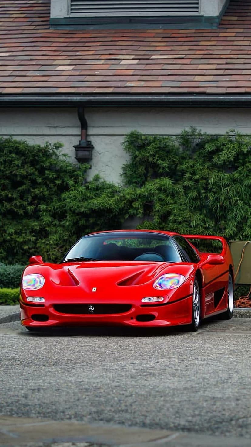 Red Ferrari F50 supercar parked on a paved driveway with dense green bushes and a tiled roof in the background.