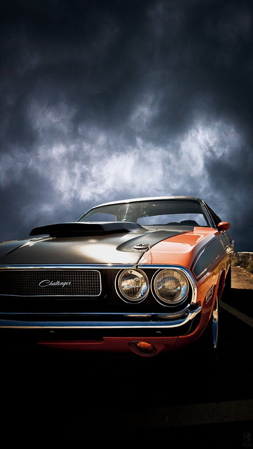 Orange Dodge Challenger muscle car with black hood scoop parked under a dark stormy sky with glowing headlights.
