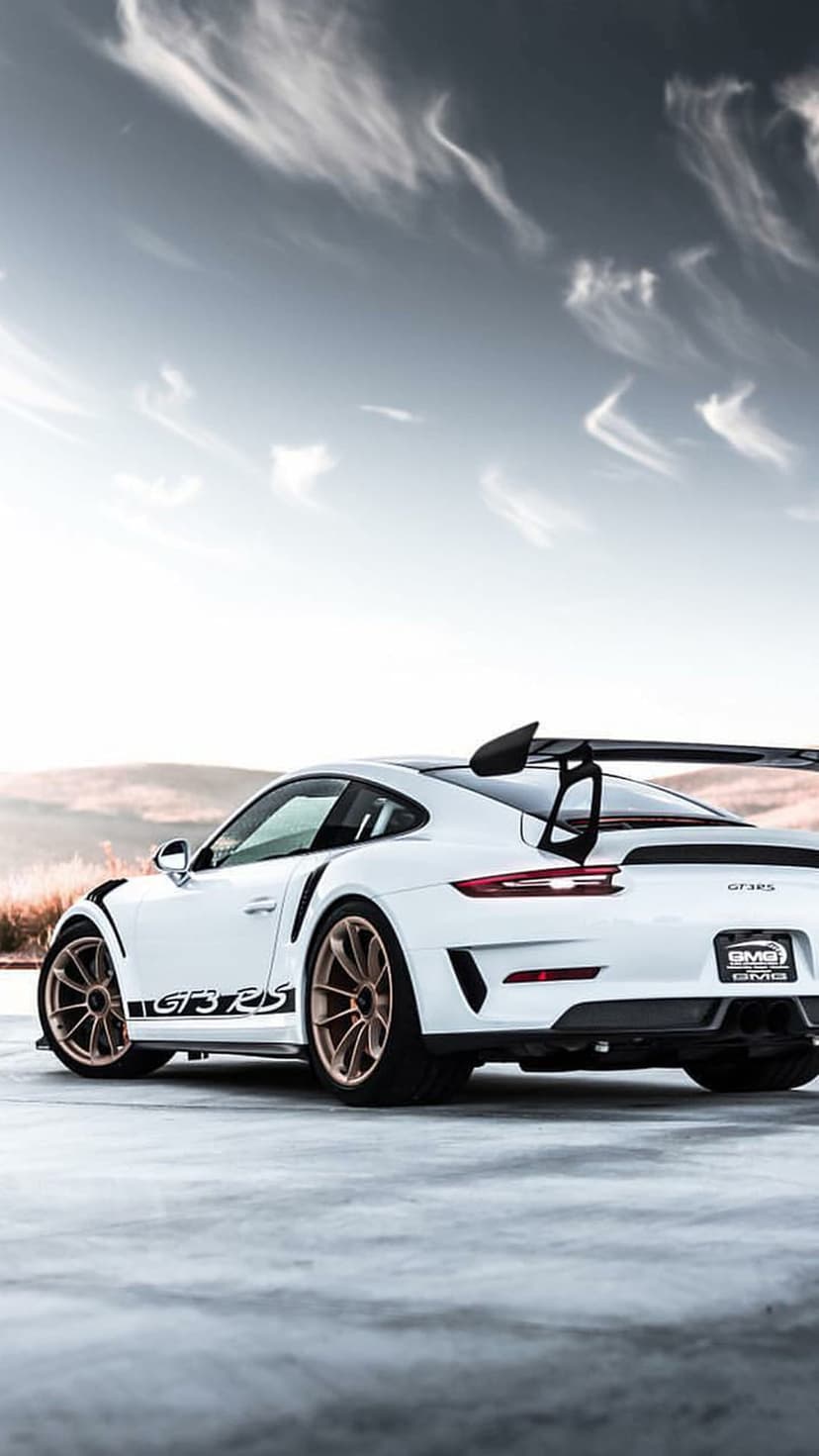 White Porsche GT3 RS showing a rear wing and gold wheels on a road under a wispy sky with hills in distance.
