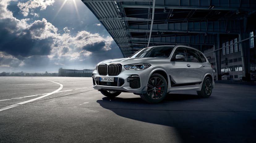 Silver BMW X5 M Performance SUV parked on an airfield tarmac under a industrial hangar with dramatic sun rays.