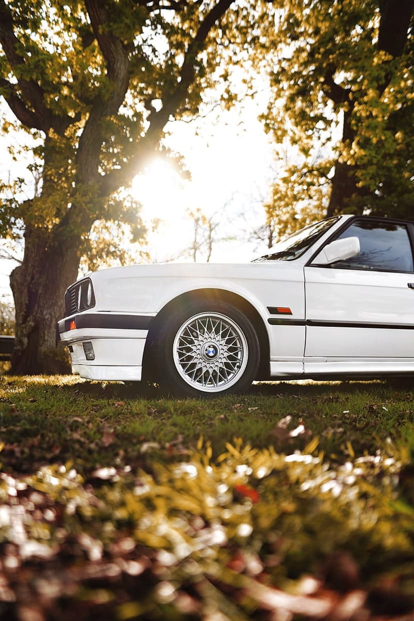 White BMW E30 convertible with BBS wheels parked under sunlit orange autumn foliage on a leaf-covered path.