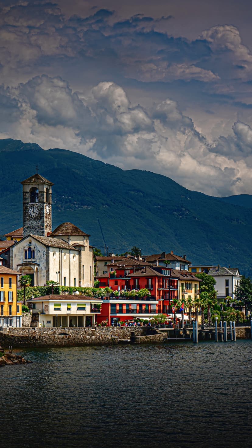 Colorful lakeside houses and a stone clock tower stand before dramatic mountains under a cloudy sky.