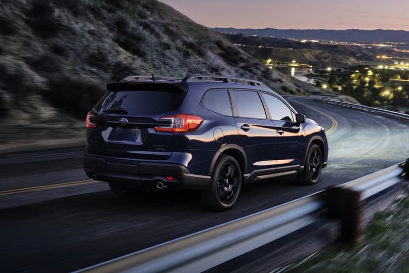 Blue Subaru Ascent driving on a winding road at dusk with city lights reflecting in the valley background.