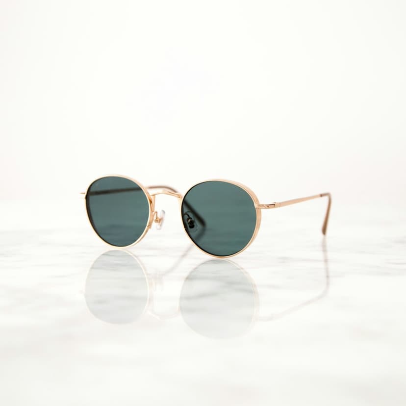 Round gold metal sunglasses with dark green lenses rest on a surface in a minimalist portrait composition.