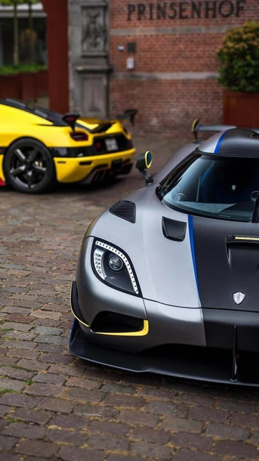 Silver and black Koenigsegg with blue stripes parked ahead of a yellow supercar on a cobblestone city street.