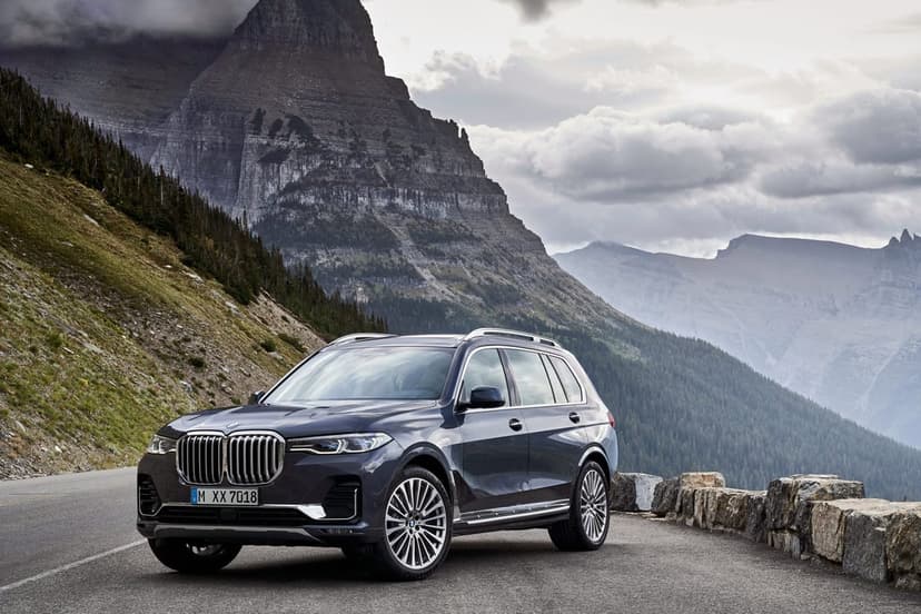 Dark blue BMW X7 parked on a mountain road with a massive rocky peak and cloudy sky in the background.