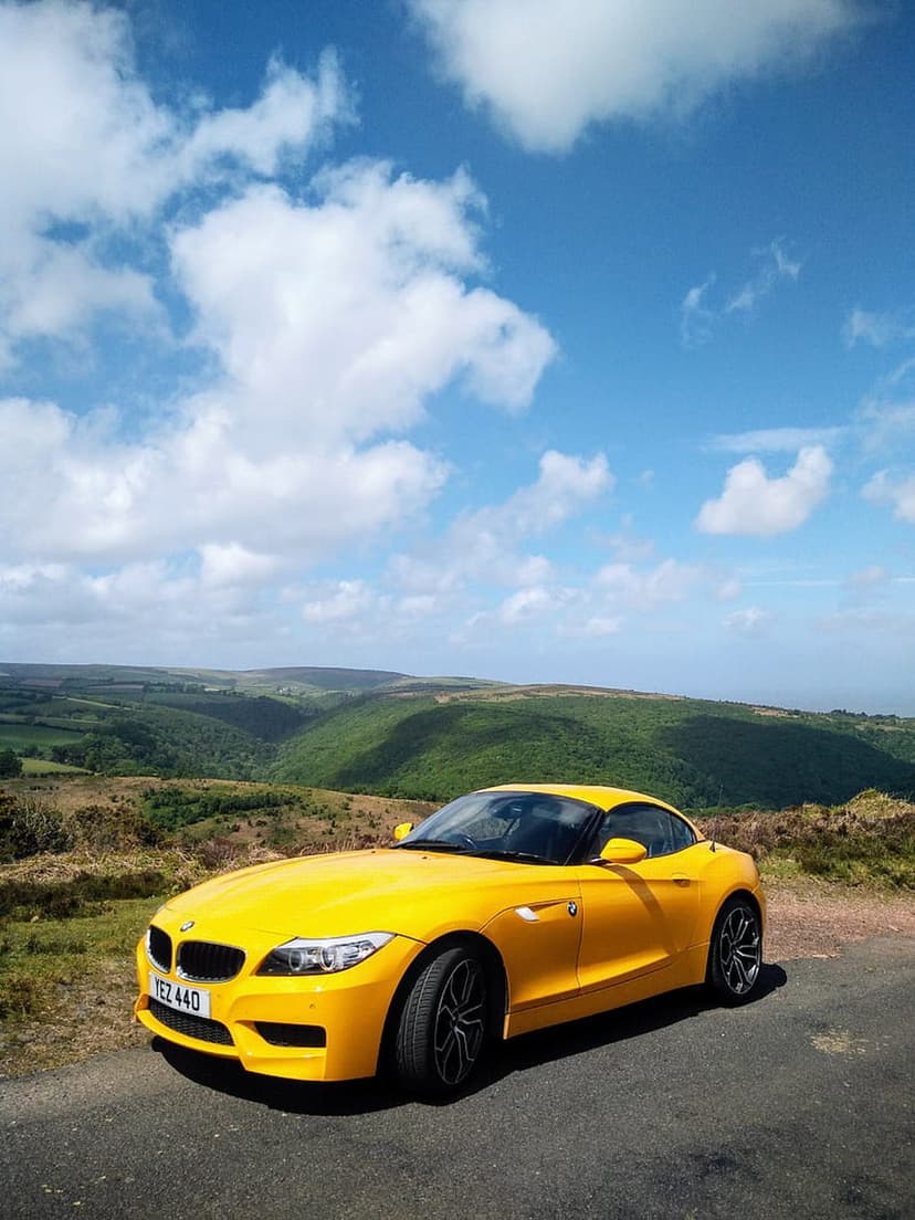 Yellow BMW Z4 convertible parked on a winding asphalt road overlooking a lush green valley under a blue sky.