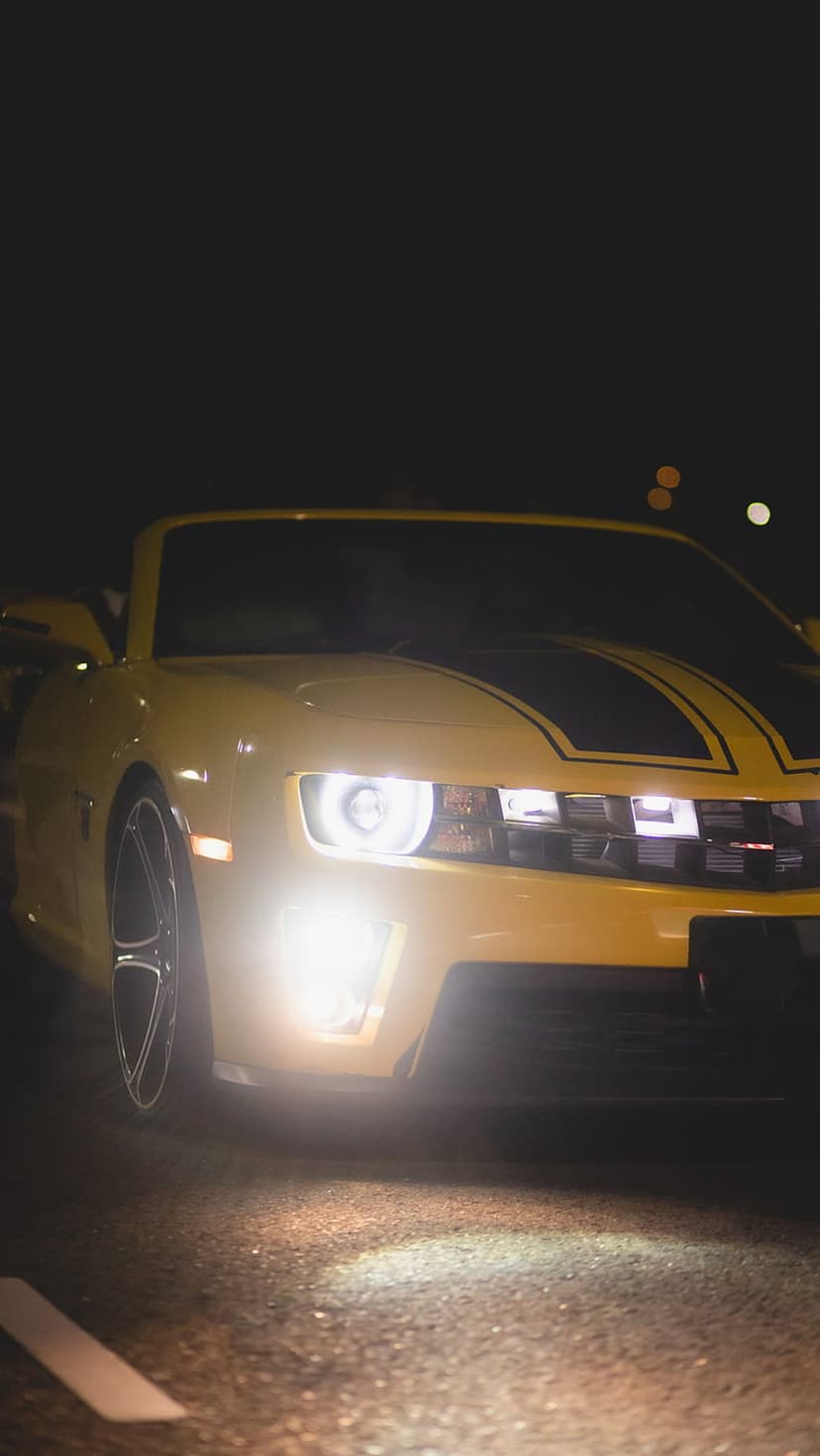 Yellow Chevrolet Camaro convertible with lit headlights and black racing stripes on a dark road at night.