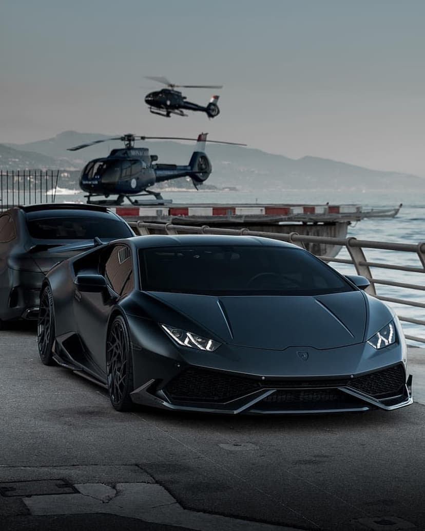 Matte black Lamborghini and luxury SUV parked by a misty sea with two helicopters flying over coastal mountains.