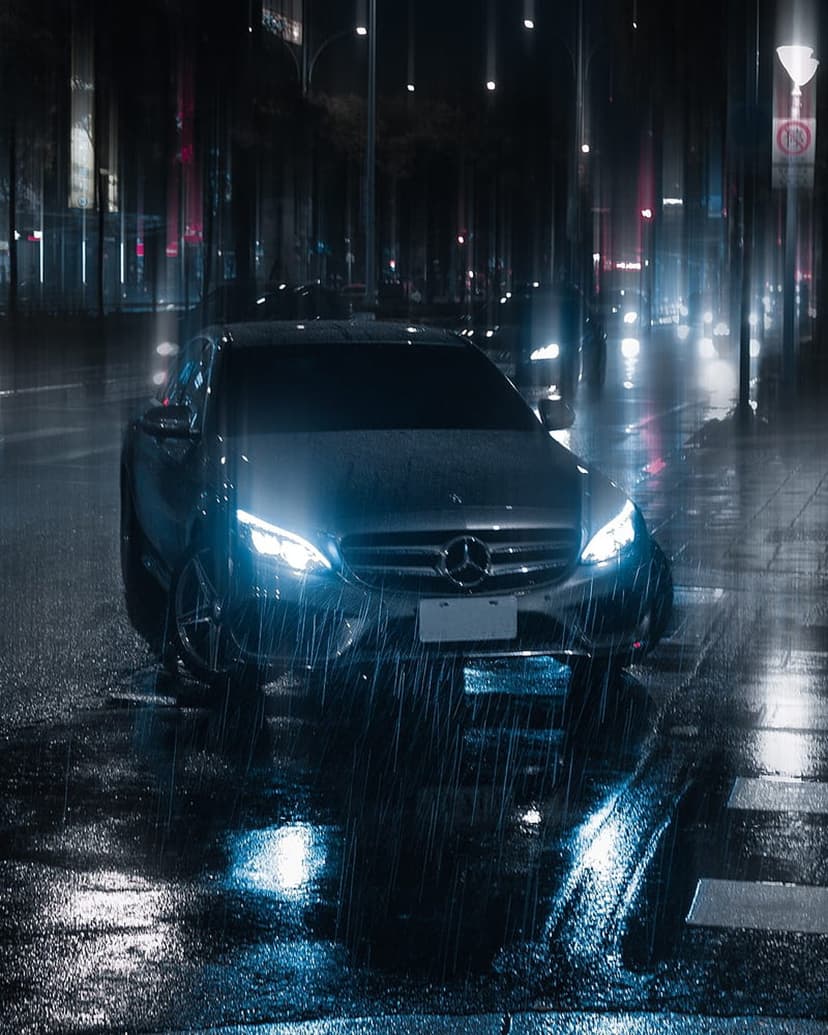 Mercedes sedan with glowing headlights on a wet city street reflecting neon lights during a rainy night.