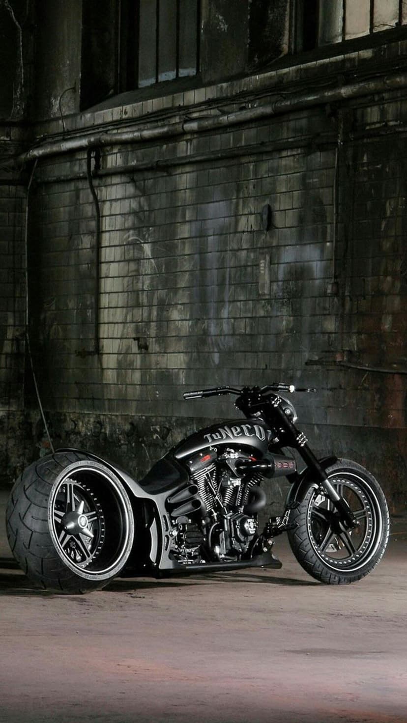 Custom black chopper motorcycle with a wide rear tire parked against a brick wall in a dark industrial garage.