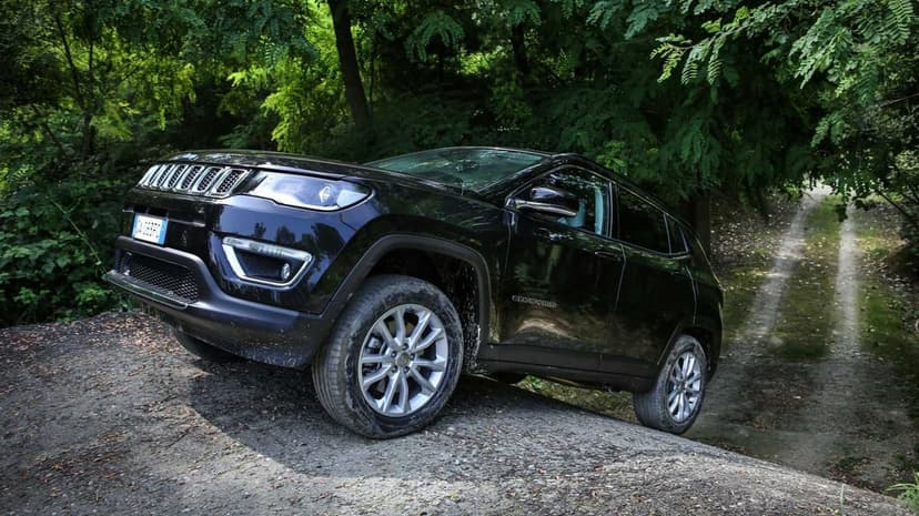 Black Jeep Compass SUV climbing a steep dirt mound on a forest trail with dense green trees in the background.