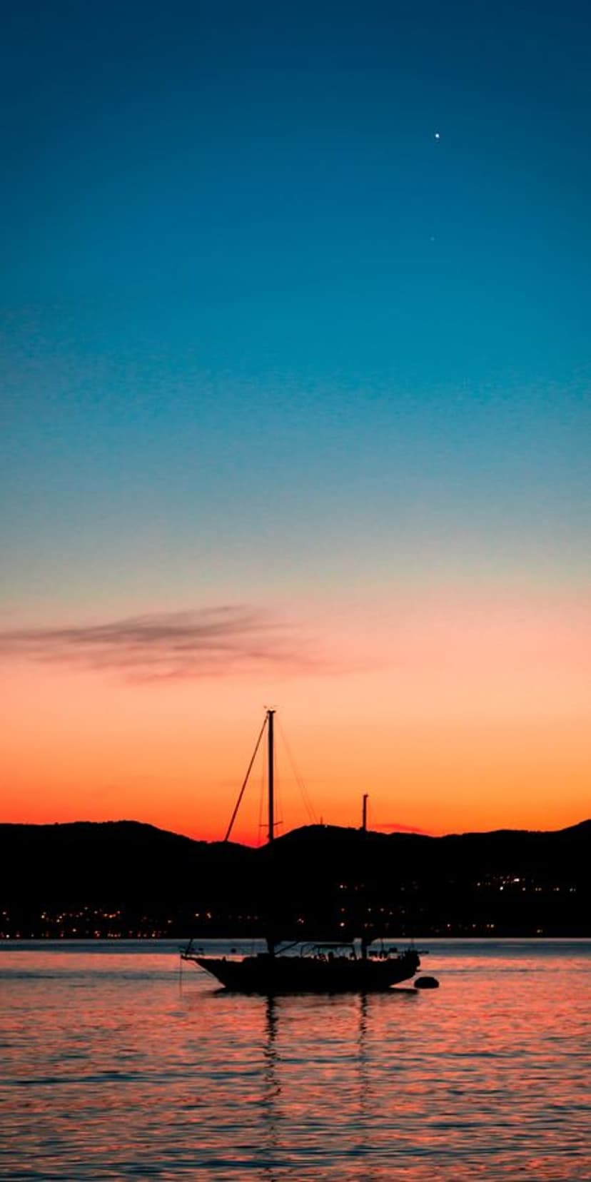 Sailboat silhouette on calm water under a red sunset sky with a single star and distant glowing city lights.