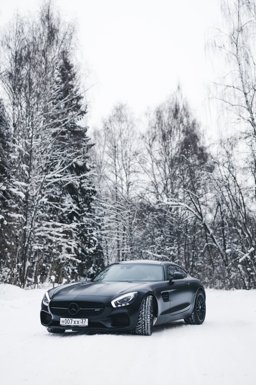 Black Mercedes AMG GT sports car with glowing headlights parked on a snowy road between winter trees.