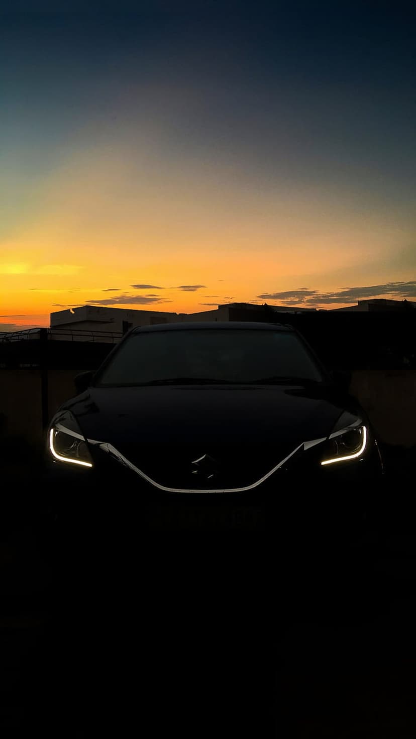 Black car headlights glowing at dusk under an orange and blue gradient sky with a faint cityscape silhouette.
