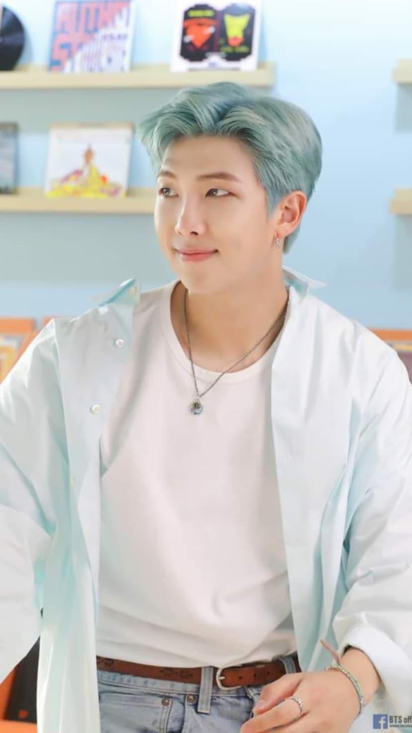 Kim Namjoon with pastel blue hair wears a denim shirt over a white tee against a blurred library background.
