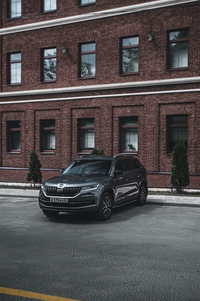 Black SUV parked on a city street in front of a red brick building with dark windows and small green trees nearby.