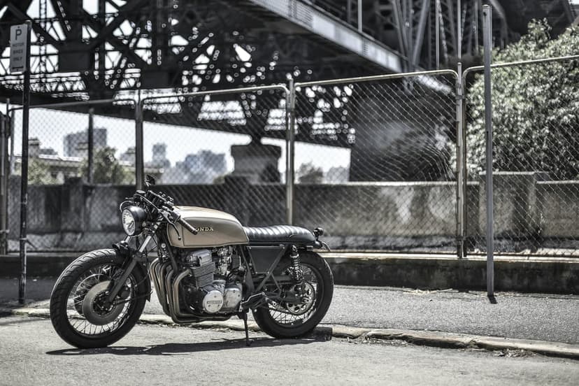 Tan and black cafe racer motorcycle parked under a massive steel bridge with an industrial city background.
