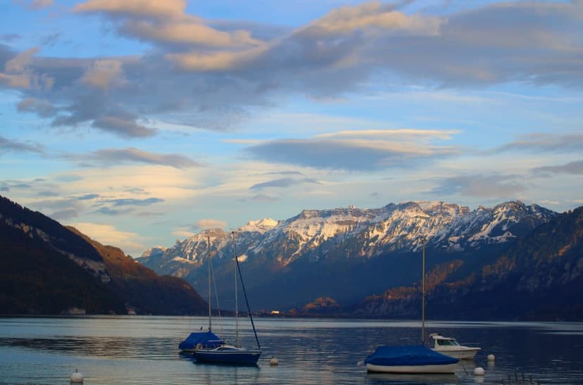 Snow-capped mountains and moored sailboats reflect in a calm lake under a golden sunset sky during winter.