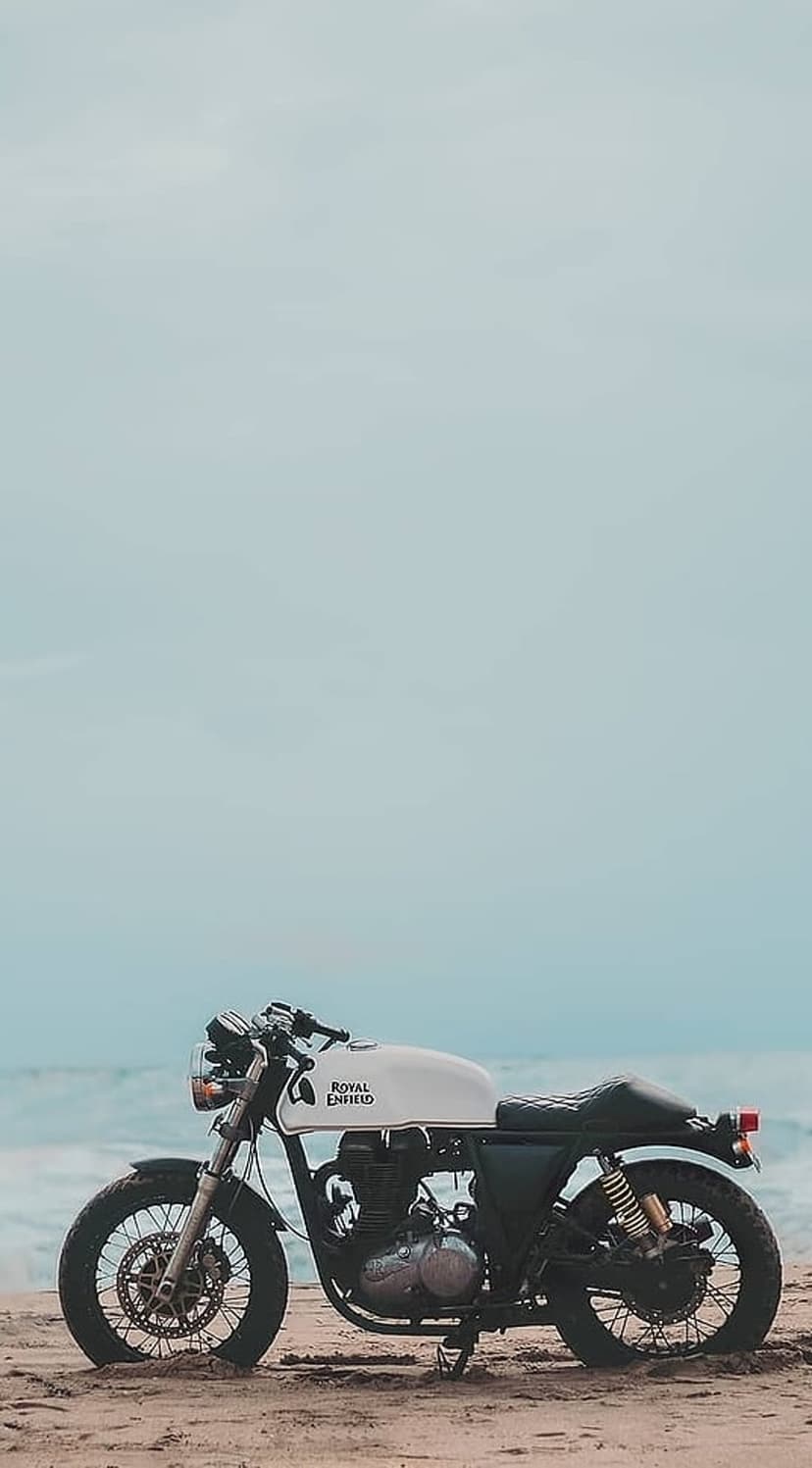 Royal Enfield motorcycle with a white fuel tank parked on a sandy beach against a blue sky and ocean horizon.