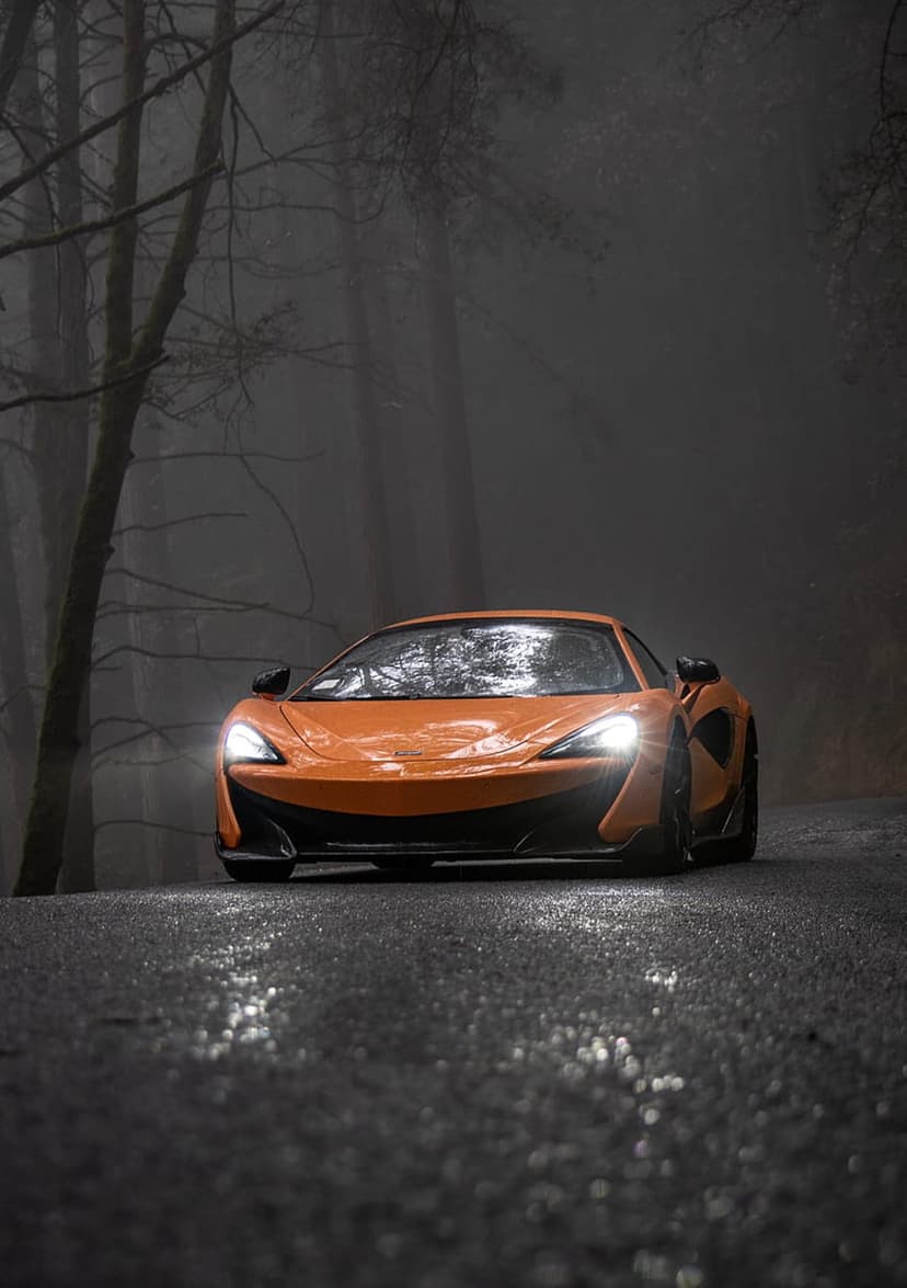 Orange McLaren sports car drives on a wet asphalt road through dense fog and dark bare trees with headlights on.