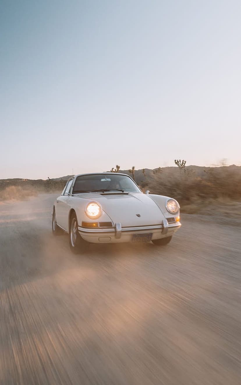 White Porsche 911 kicks up dust on a desert road with glowing headlights under a hazy golden sunset sky.