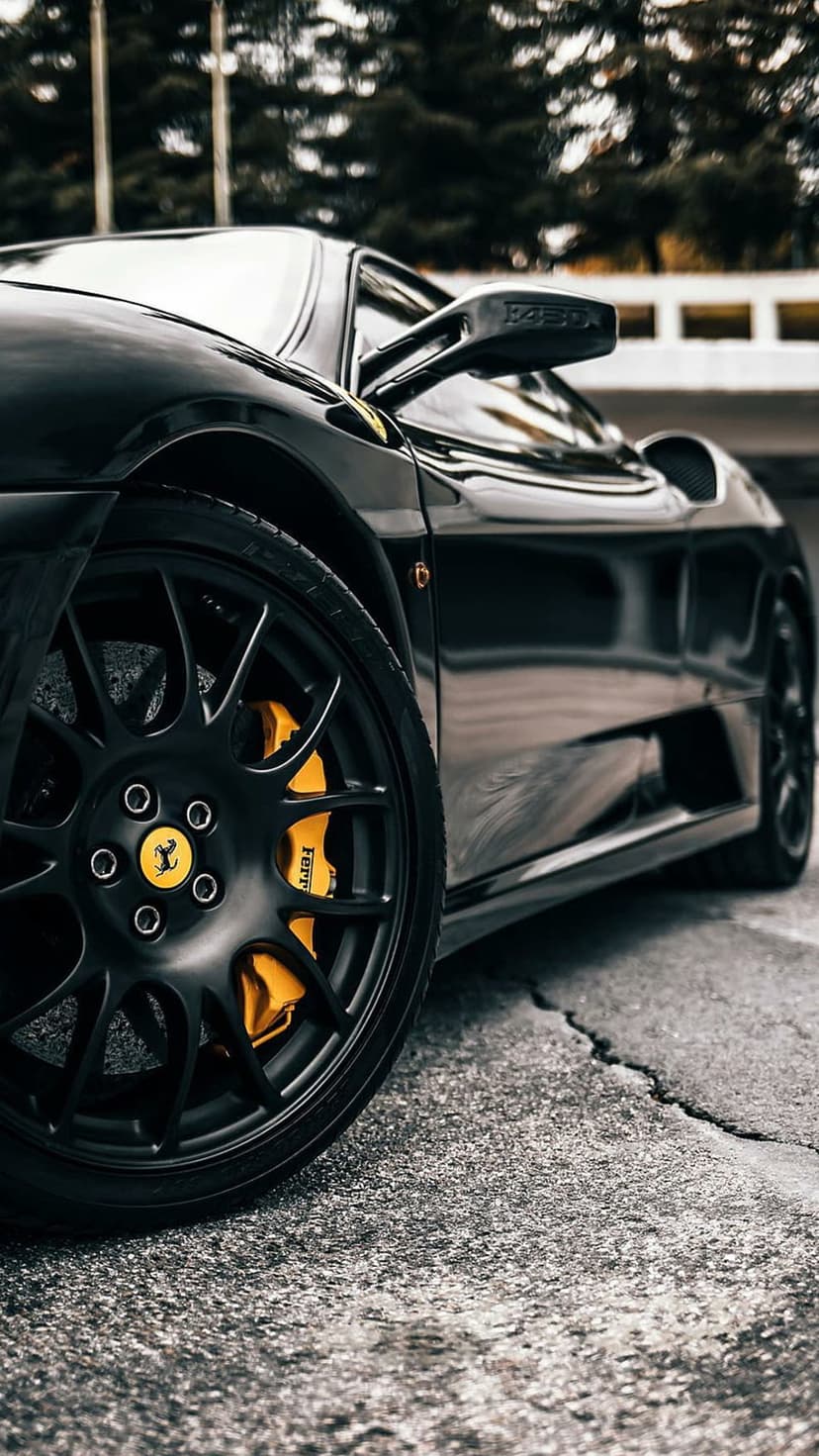 Ferrari F430 black alloy wheel showcasing a bright yellow brake caliper and reflective bodywork paint.