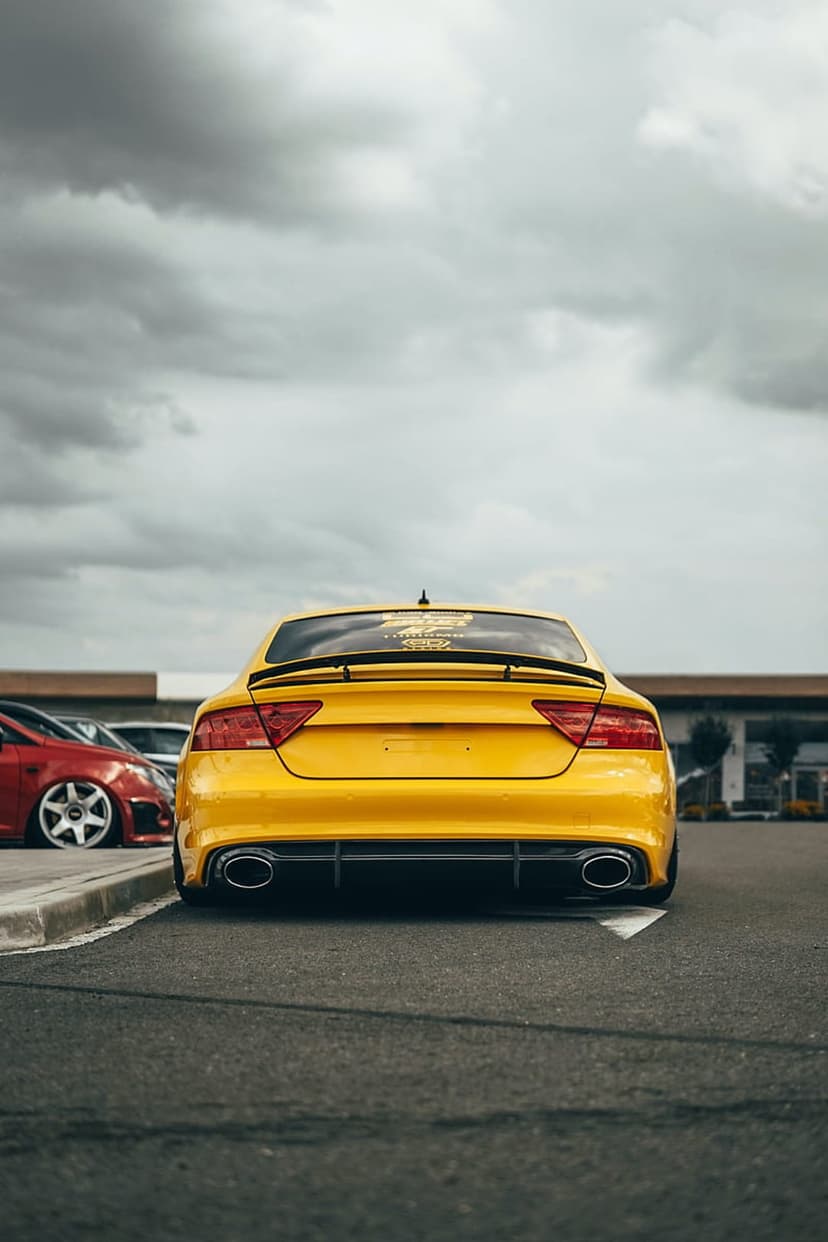 Yellow Audi A7 with a large rear spoiler and diffuser parked on dark asphalt under a heavy, overcast cloudy sky.