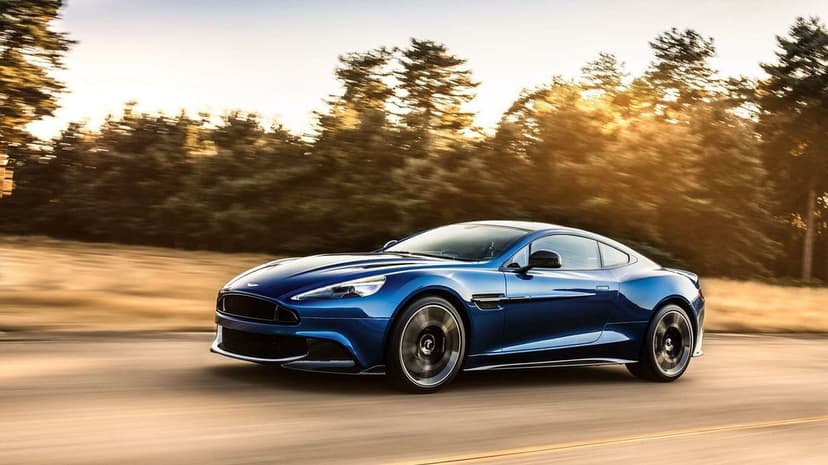 Blue Aston Martin Vanquish driving fast on a sunlit road through a forest with golden motion blur.