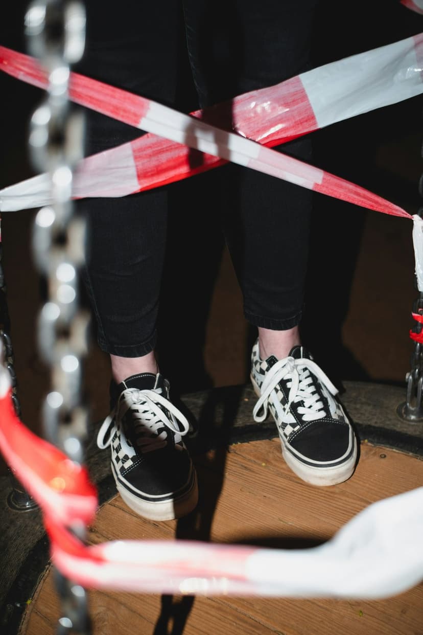 Checkerboard sneakers and black trousers stand behind a red and white caution tape with a metal chain nearby.