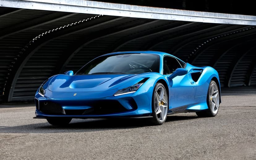Blue Ferrari F8 Tributo parked on asphalt under a curved metal industrial canopy with bright daylight highlights.