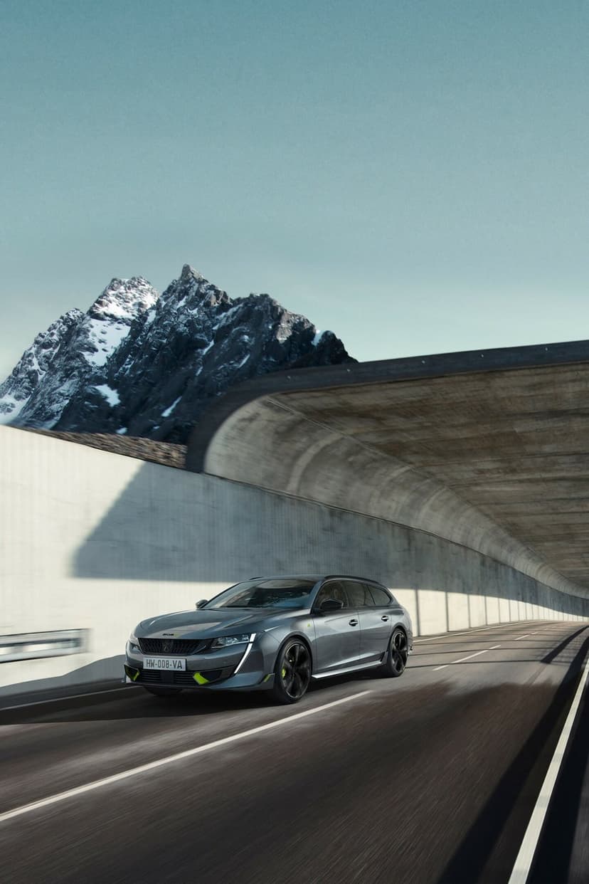 Grey sports wagon driving on a mountain road beside a concrete tunnel under a clear sky with snowy peaks.