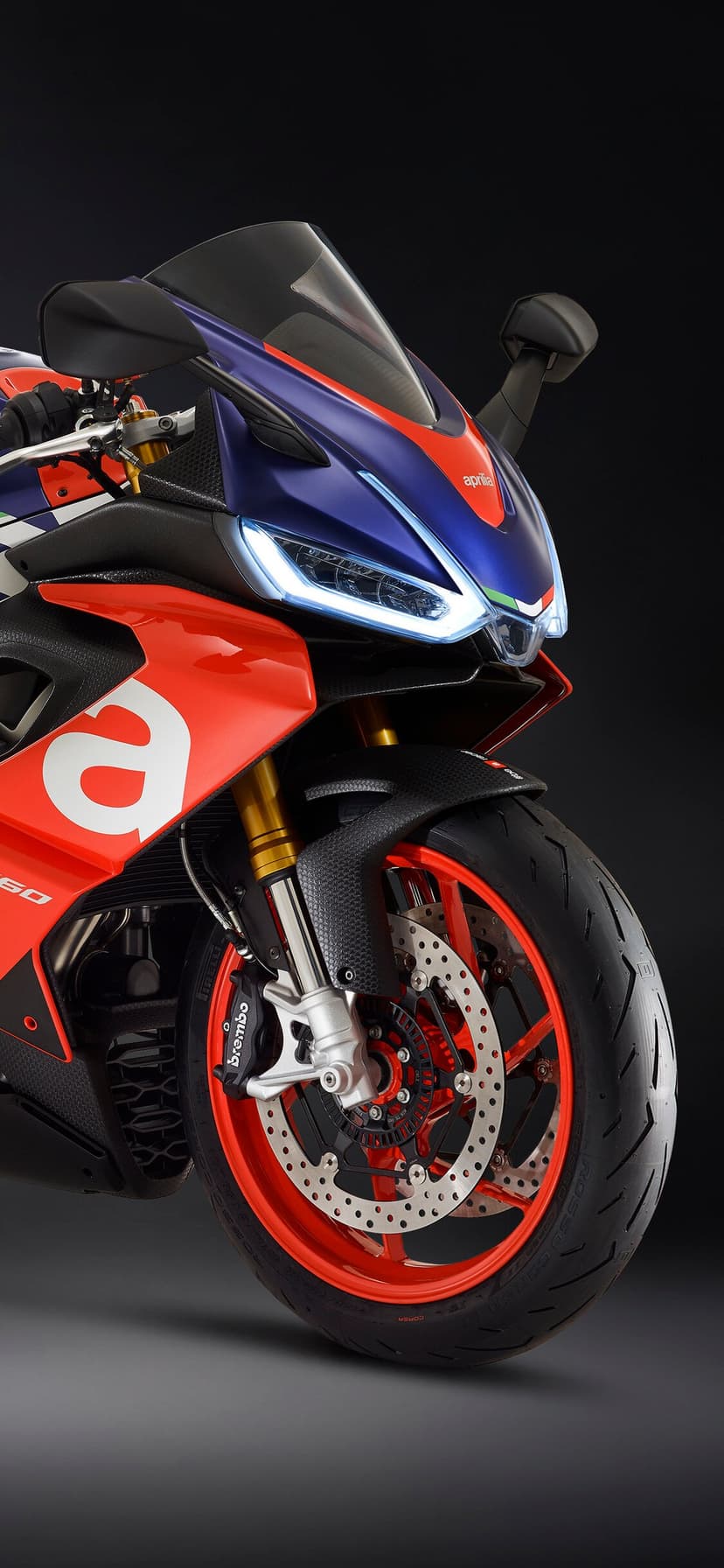 Close-up of an Aprilia motorcycle front with glowing LED headlights, carbon fiber fender, and red wheels.