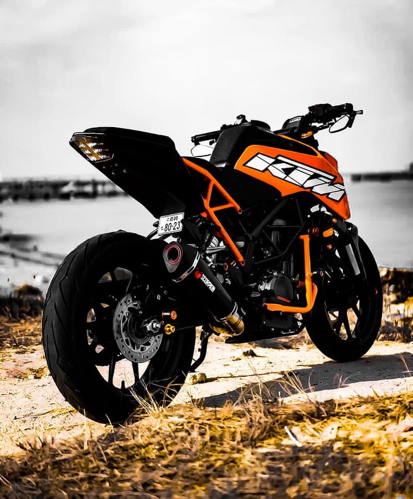 Orange KTM motorcycle parked on a sandy beach viewed from the rear under a dark, dramatic overcast sky.