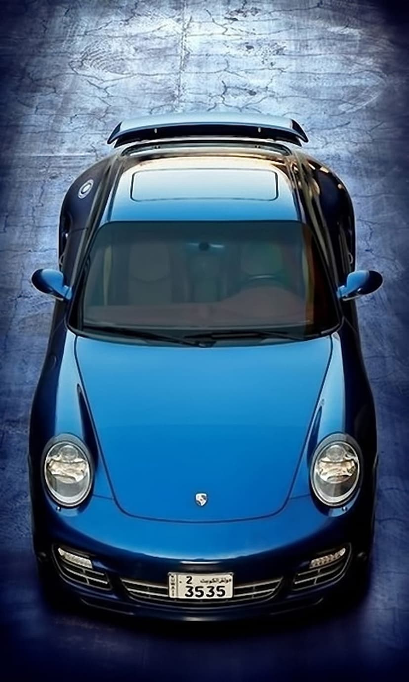 Blue Porsche 911 sports car viewed from above on a dark textured floor with an Arabic script license plate.