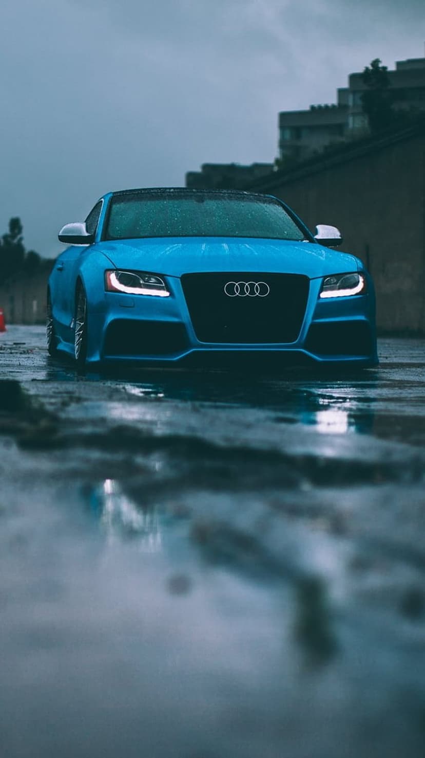 Blue Audi S5 convertible parked on a wet, reflective urban street with raindrops visible on the metallic body.
